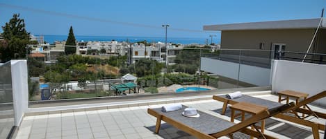 A rooftop terrace with wooden loungers, a pool, and a view of the sea and buildings.