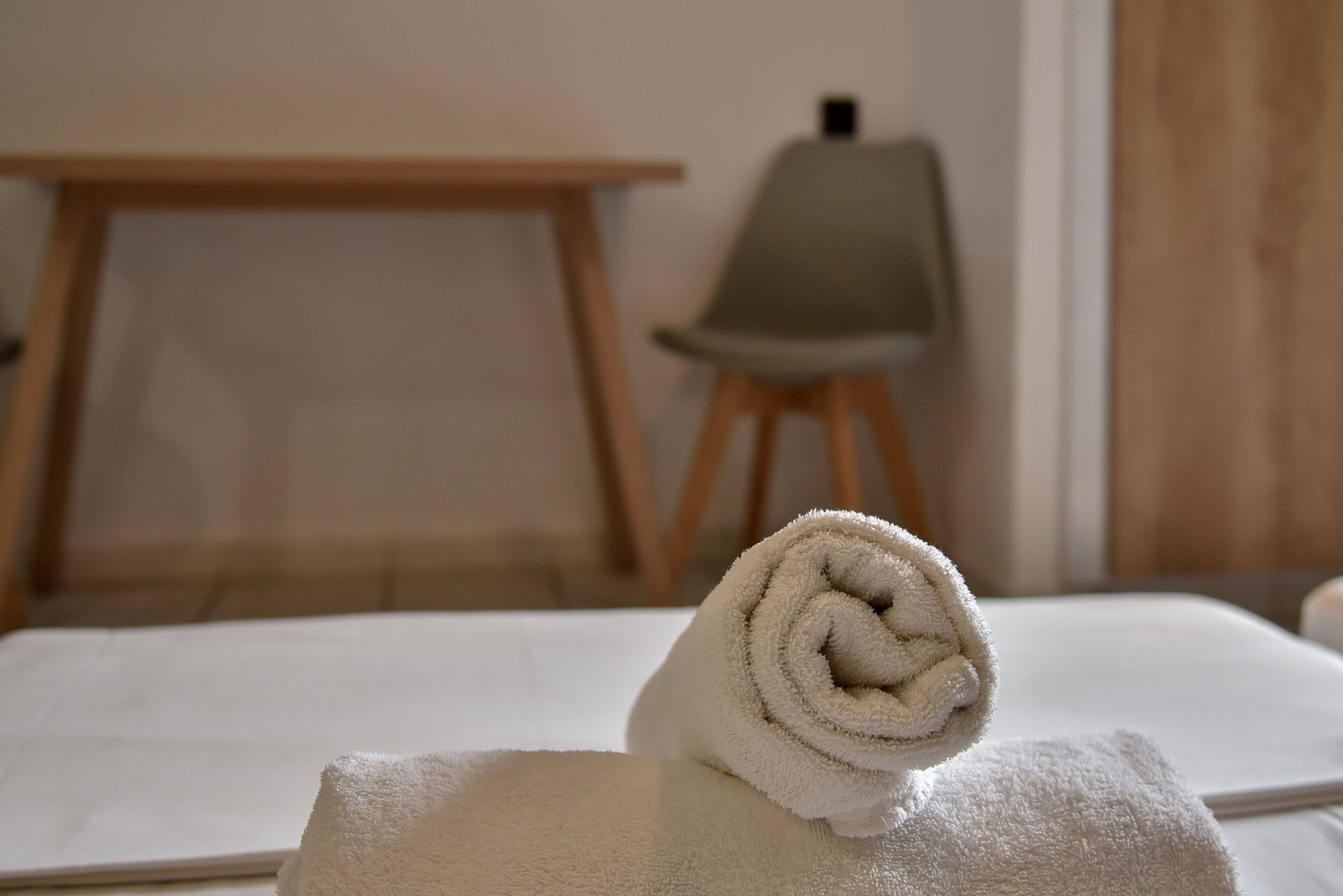 Una cama bien hecha con una toalla blanca enrollada encima, una silla de madera y una mesita de noche en una habitación con pared lisa y una puerta.