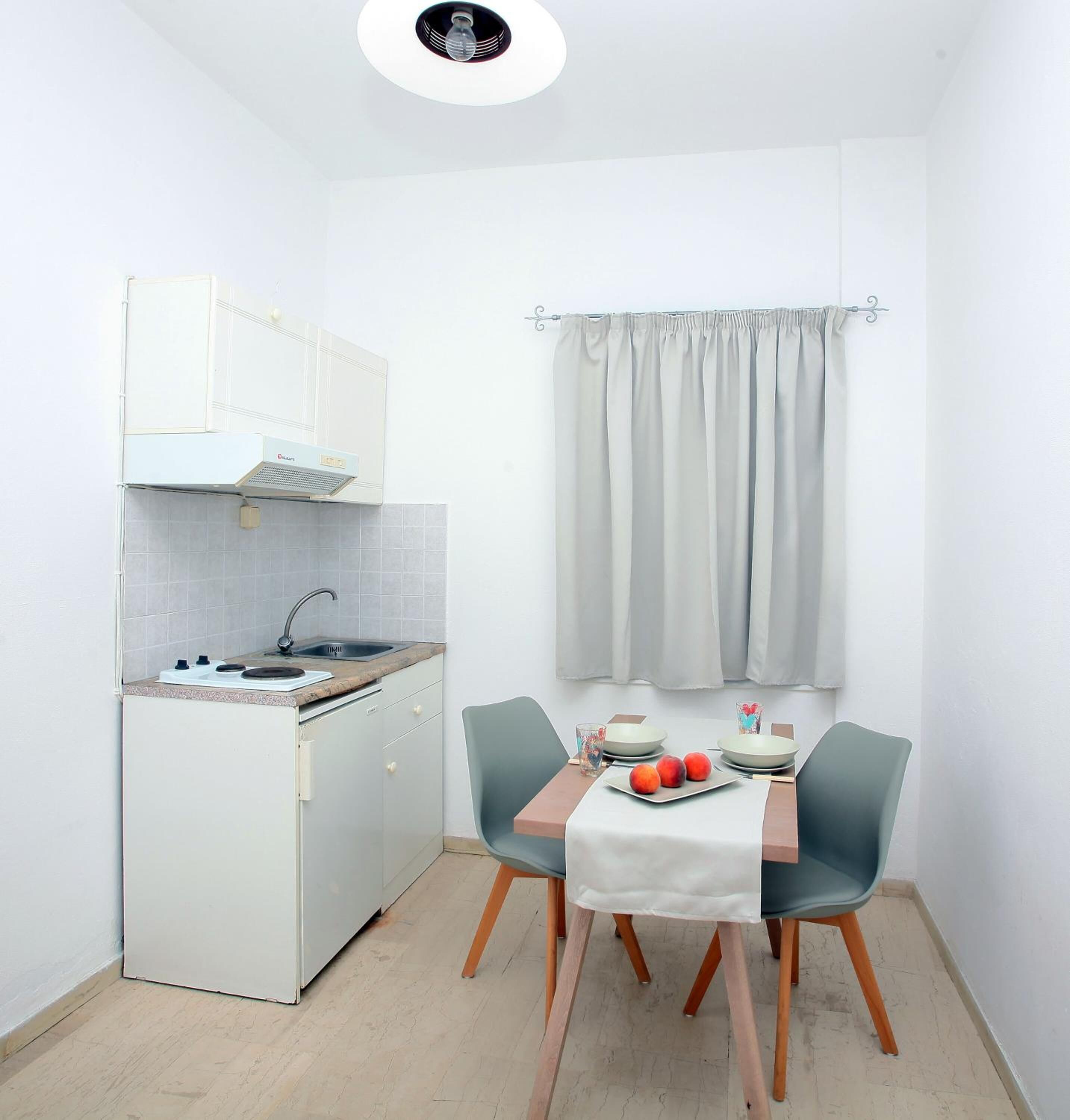 A compact kitchen with a stove, sink, and white cabinets. There is a dining table with two chairs and a small window with curtains.