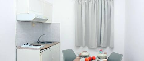 A compact kitchen with a stove, sink, and white cabinets. There is a dining table with two chairs and a small window with curtains.
