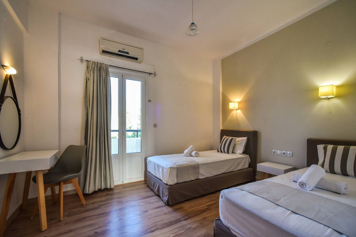 A hotel room with a desk, two beds, a chair, and a balcony.