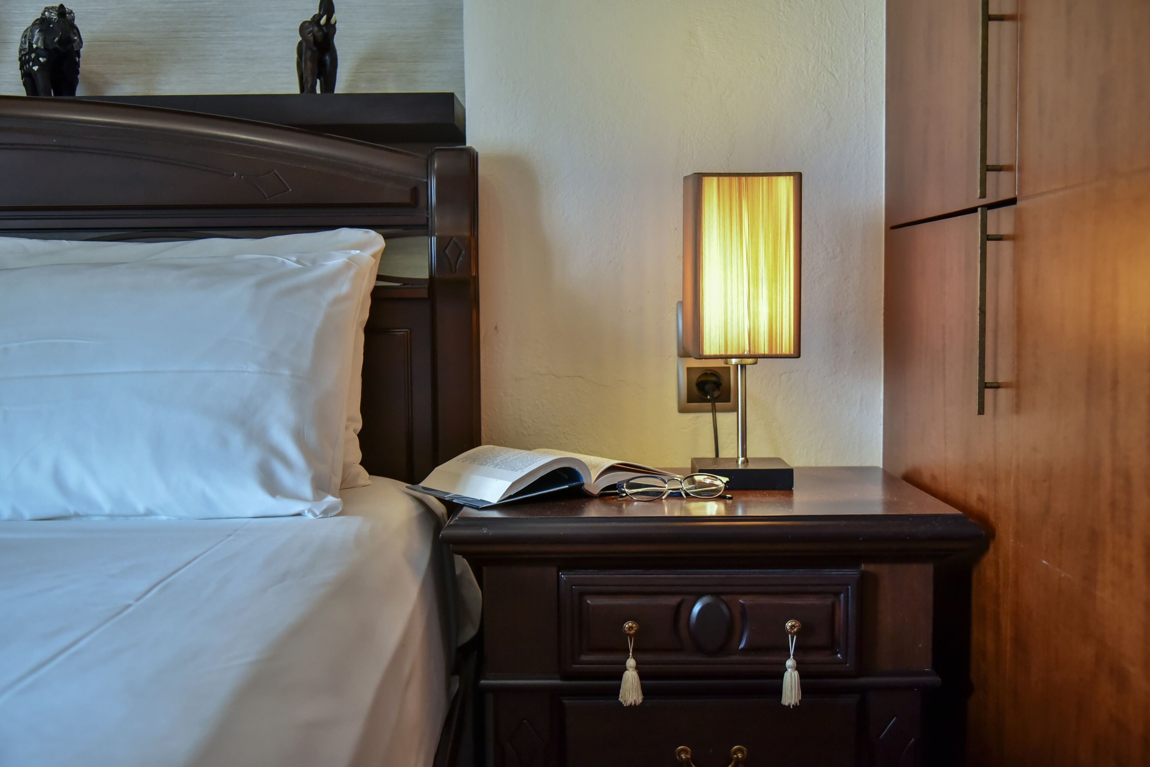 A neatly made bed with white linens, a bedside table with a lamp, and a small statue on the headboard.