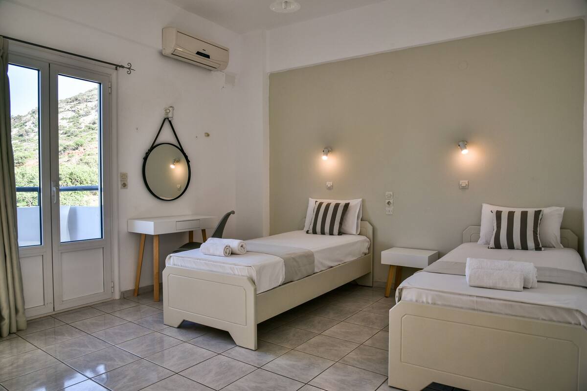 A hotel room with two beds, a small bedside table, a mirror, and a window with curtains.