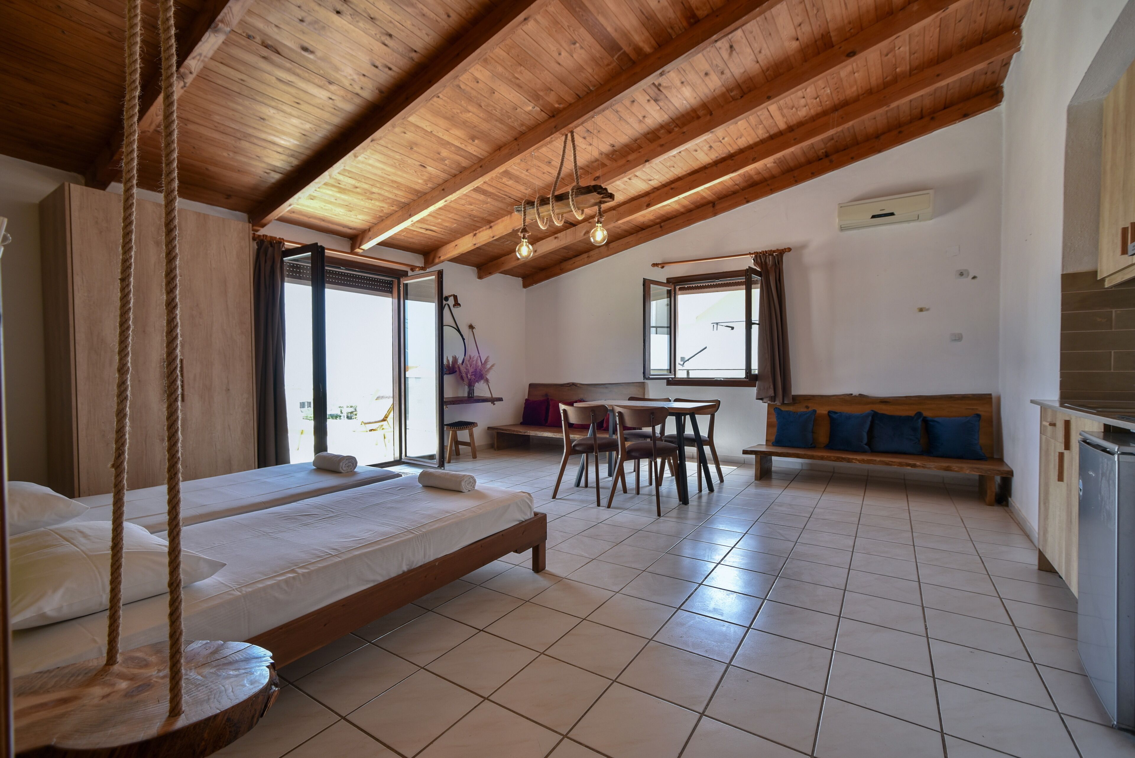 Un'ampia stanza con soffitto in legno a vista, un letto con altalena, una zona pranzo con tavolo e sedie, e una panca con cuscini.