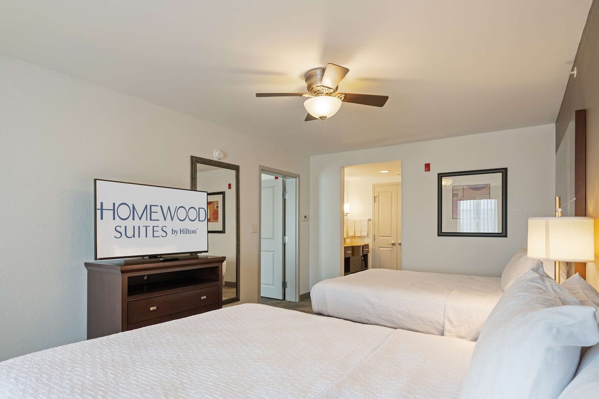 A hotel room with two beds, a ceiling fan, a TV displaying 'HOMEWOOD SUITES by Hilton', a mirror, and a door leading to a bathroom.