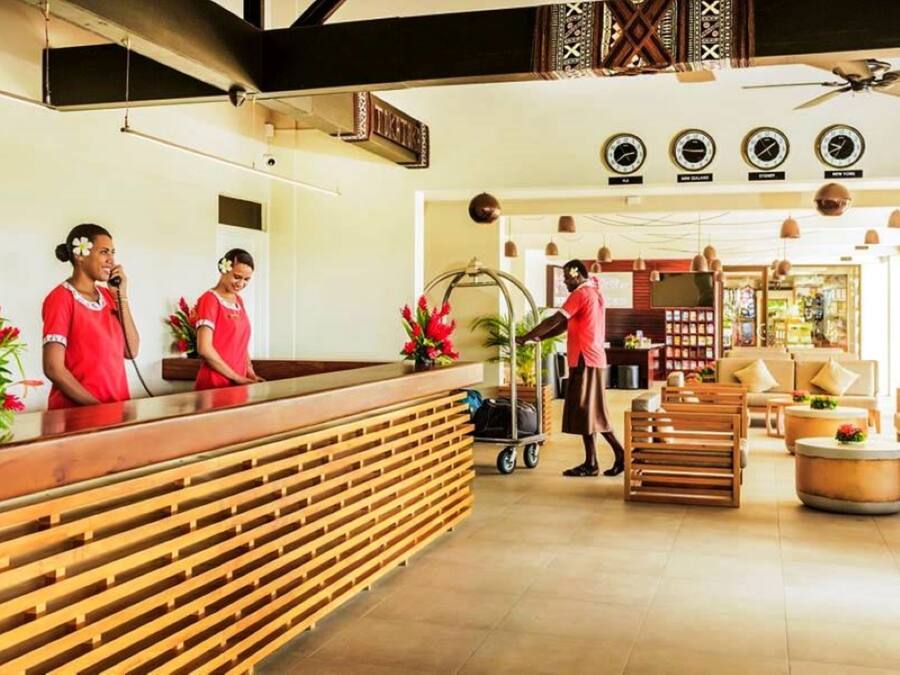 Un hall d’accueil d’hôtel avec un comptoir de réception en bois, un espace d’attente doté de canapés et d’une table basse, et un ventilateur de plafond.