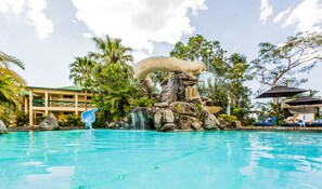 A swimming pool with a rock waterfall feature, surrounded by palm trees and a building with a green roof. - Tokatoka Resort Hotel (Nadi)