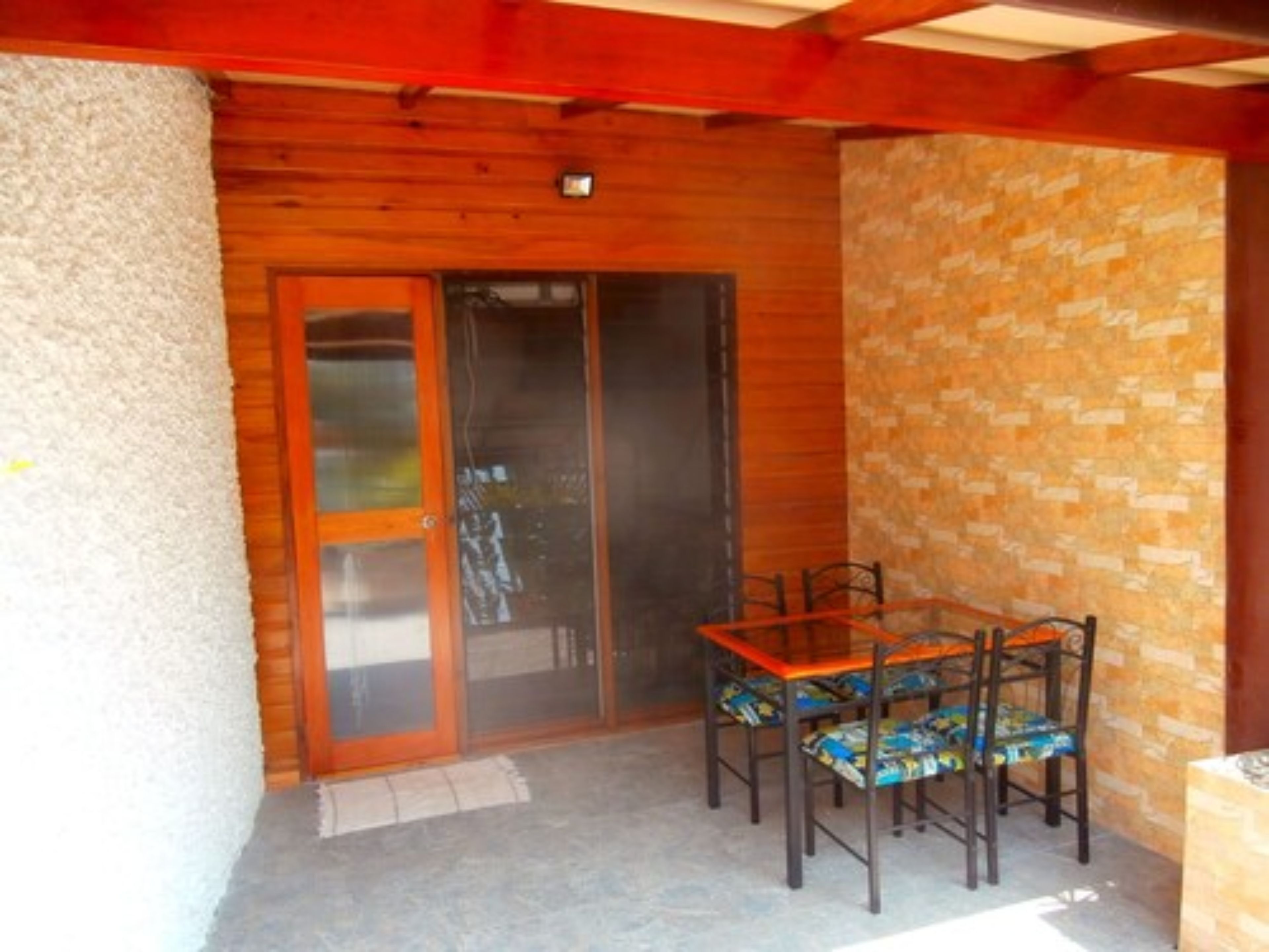 Patio con estructura de madera, una mesa, sillas, puerta corrediza de vidrio y pared con diseño de piedra.