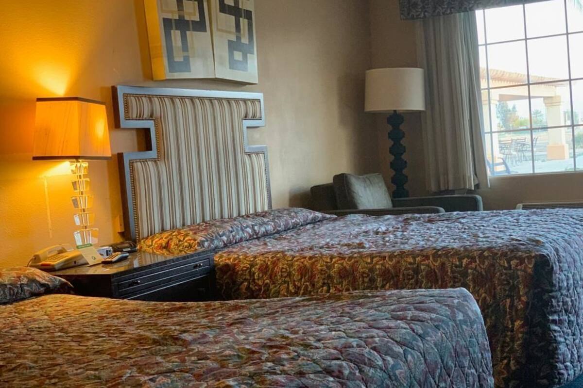 A hotel room with two beds, a nightstand with a lamp, a wall-mounted TV, and a window with curtains.