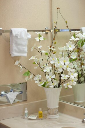 Une salle de bain avec un vase blanc contenant des fleurs blanches, une serviette suspendue à un porte-serviettes et des articles de toilette posés sur le comptoir.