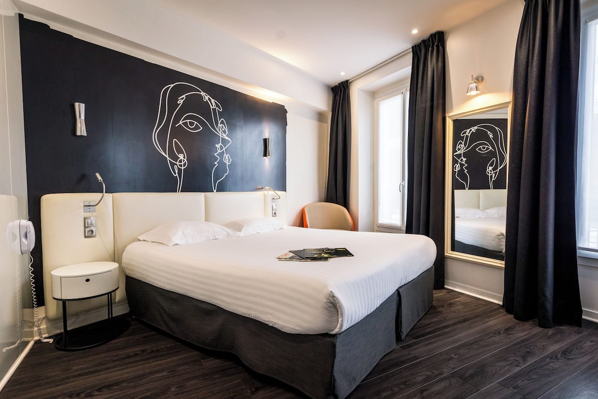 A hotel room with a large bed, a bedside table, a chair, a mirror, and a wall with a line drawing.