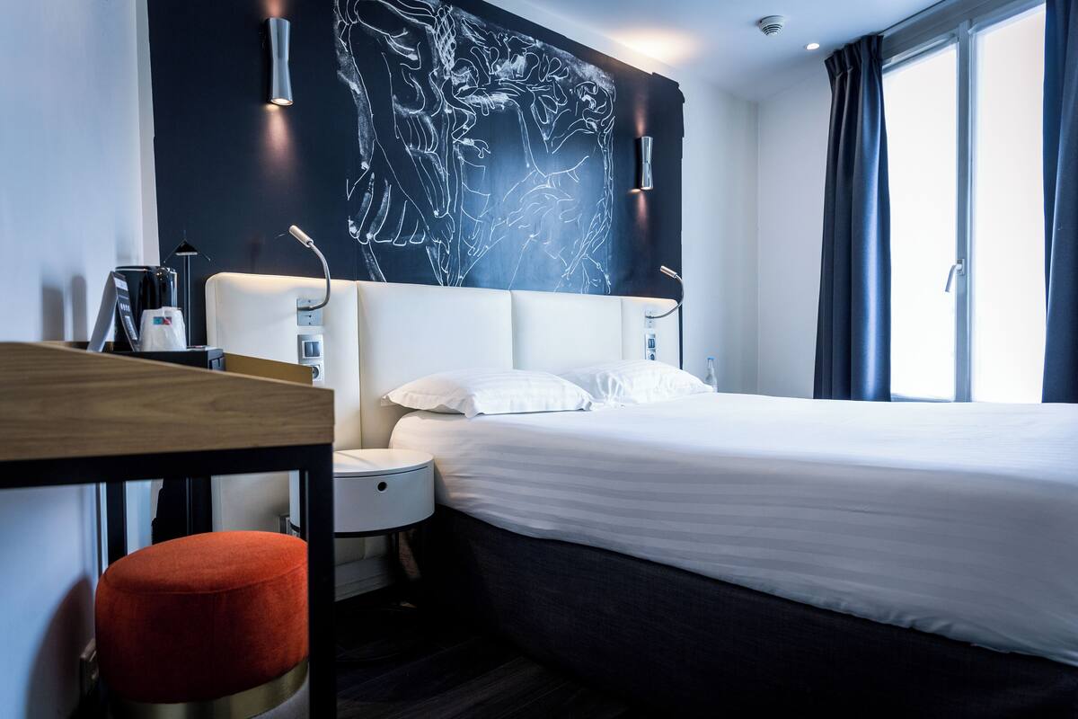 A modern hotel room with a large bed, a desk, a chair, and a wall-mounted artwork.