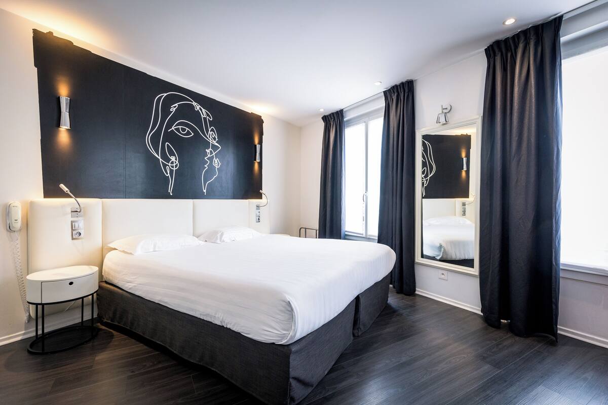 A modern hotel room with a large bed, a bedside table, a wall-mounted light, and a large mirror.