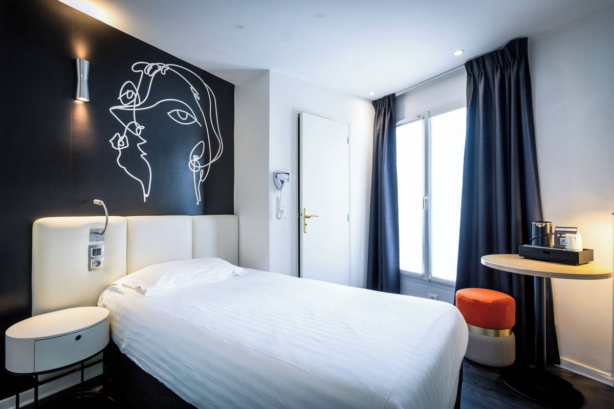 A modern hotel room with a large bed, a bedside table, a wall-mounted light, a small round table with a tray, and a window with curtains.