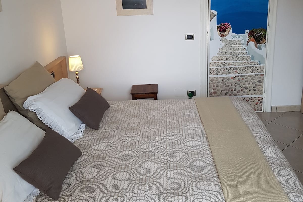 A bedroom with a bed, pillows, a nightstand, a lamp, a painting, and a view of a staircase leading to a balcony with flowers.