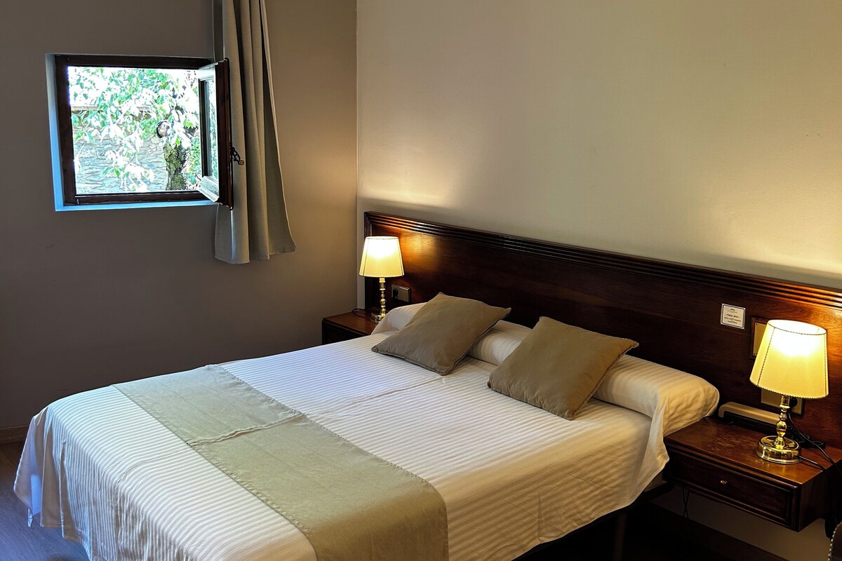 A neatly made bed with two pillows, a bedside table with a lamp, and a window with curtains.