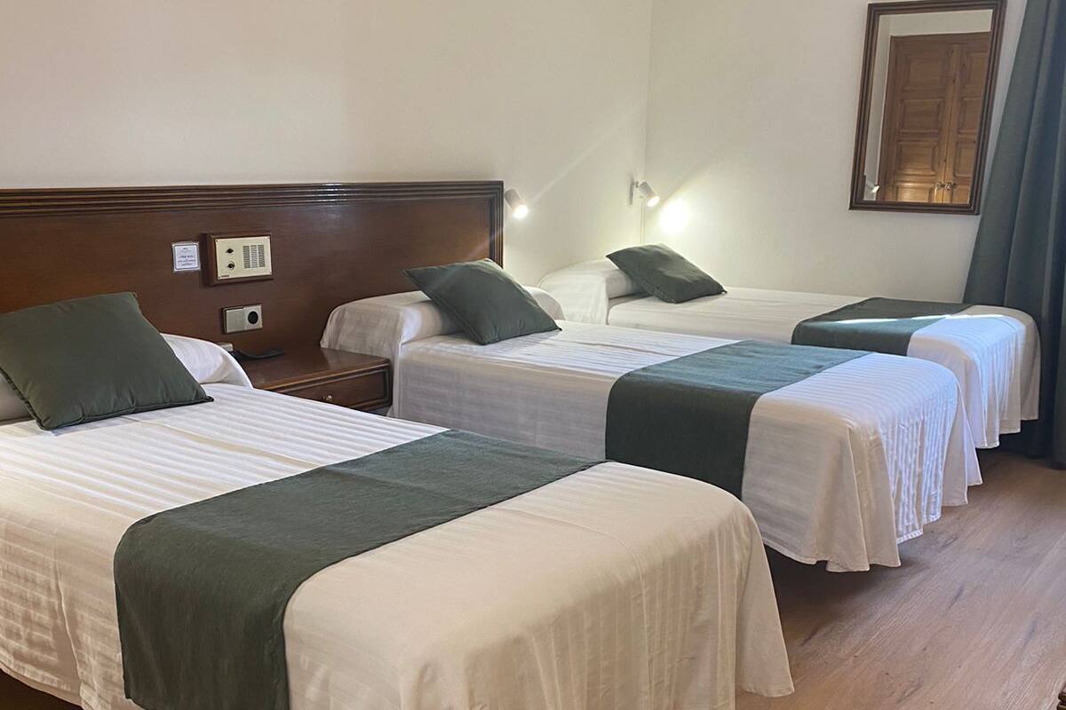 Hotellihuoneessa on kolme erillistä sänkyä, joissa kaikissa on vihreä päiväpeite ja valkoiset vuodevaatteet. Vasemmanpuoleisessa sängyssä on puinen pääty, ja sen yläpuolella seinään on kiinnitetty ohjauspaneeli.