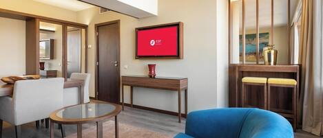 Une chambre d’hôtel moderne avec un fauteuil bleu, une table basse en verre, un bureau en bois et un téléviseur affichant le logo de l’hôtel.