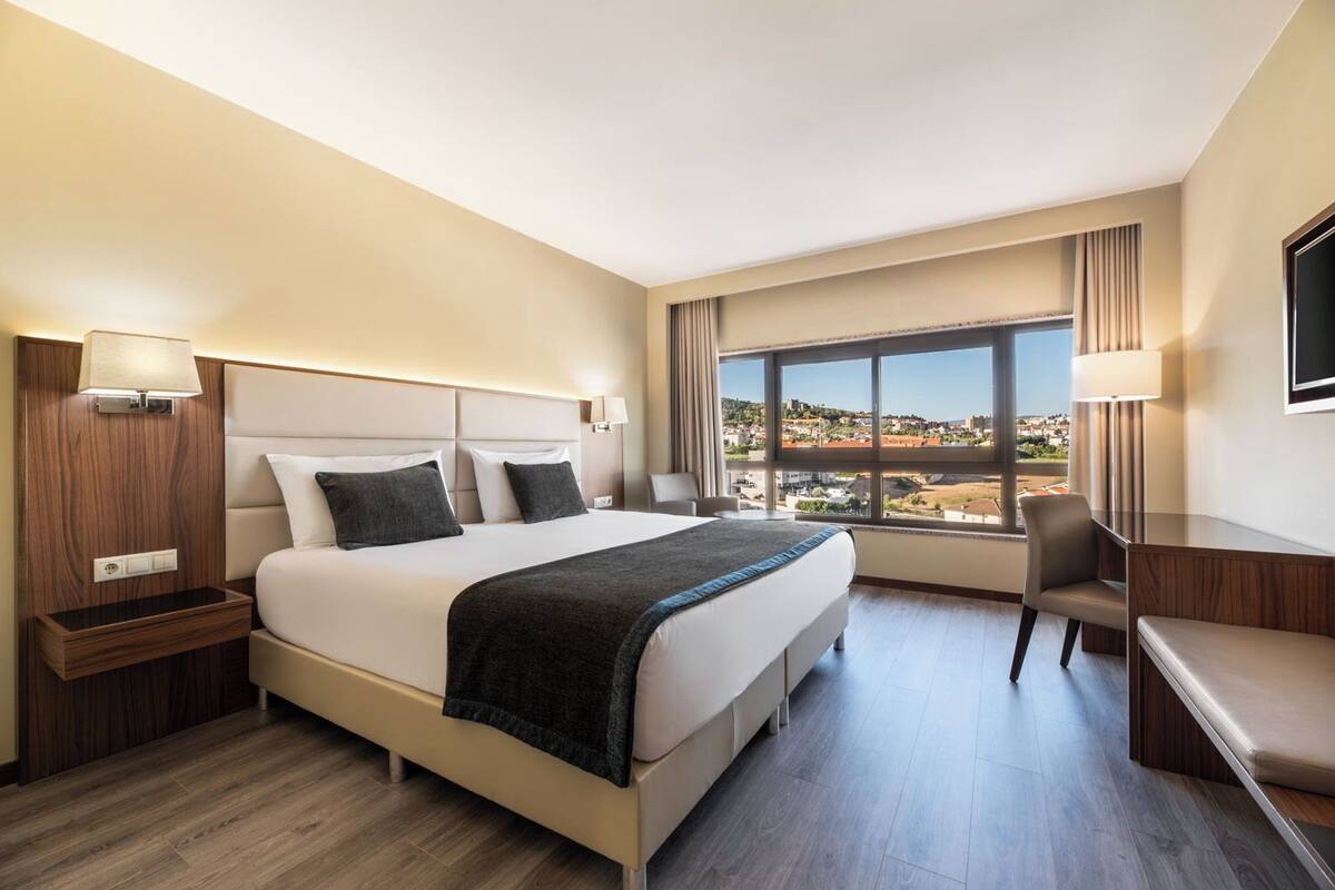 A hotel room with a large bed, a desk, a chair, a TV, and a window with a city view.