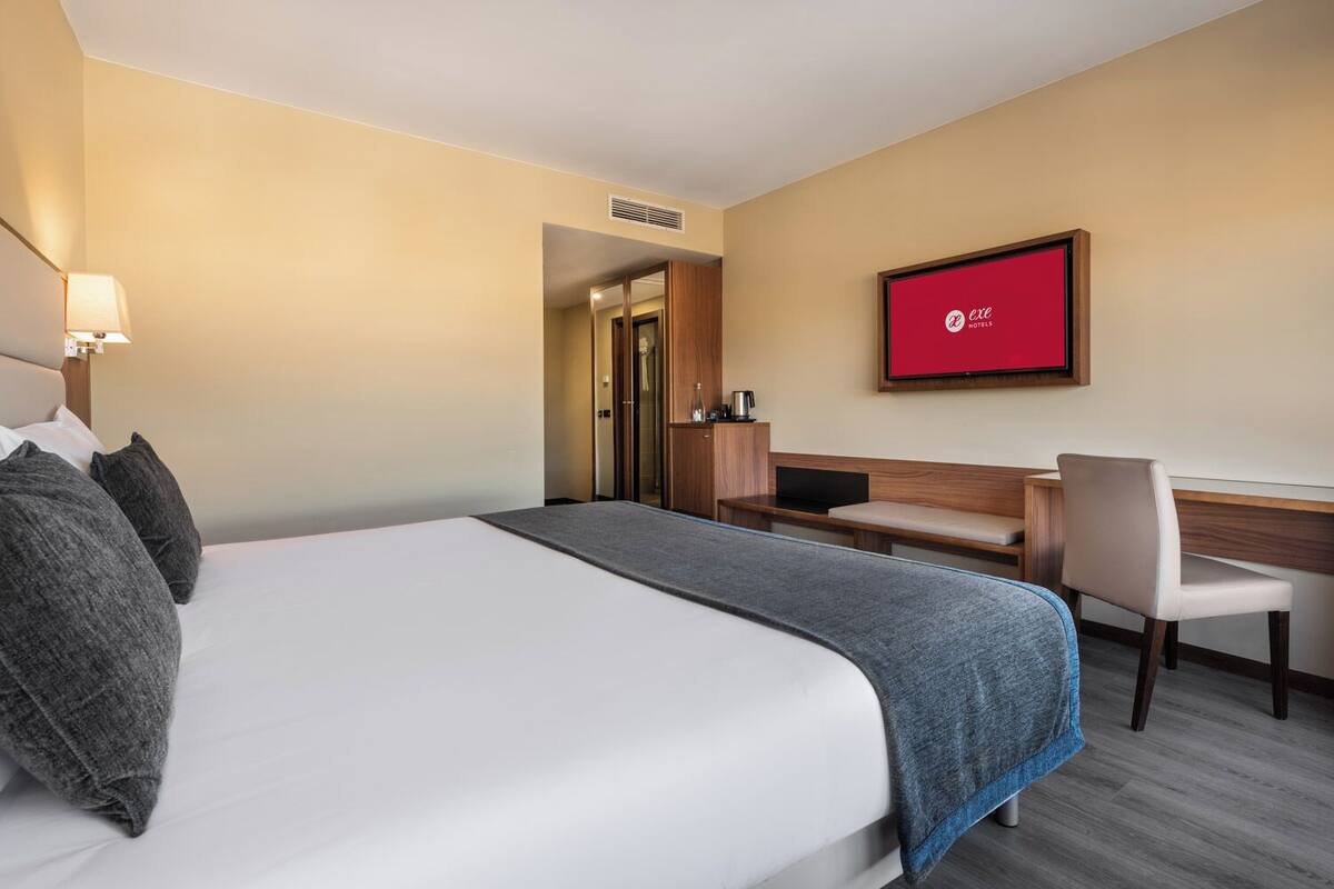 A hotel room with a bed, a desk, a chair, a TV, and a minibar.