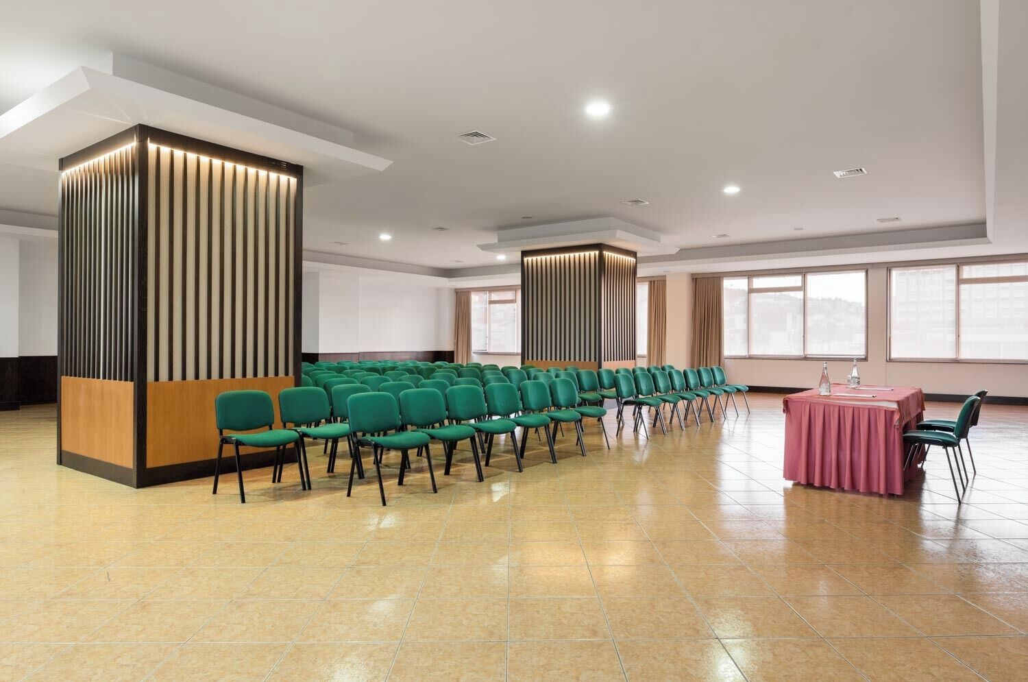 Un salón de conferencias con hileras de sillas verdes, una mesa rosa y modernas lámparas verticales.