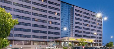 A multi-story hotel with a modern facade, featuring a prominent glass section and a ground-level entrance with a sign for "HOTEL TURISMO LÁZARO".