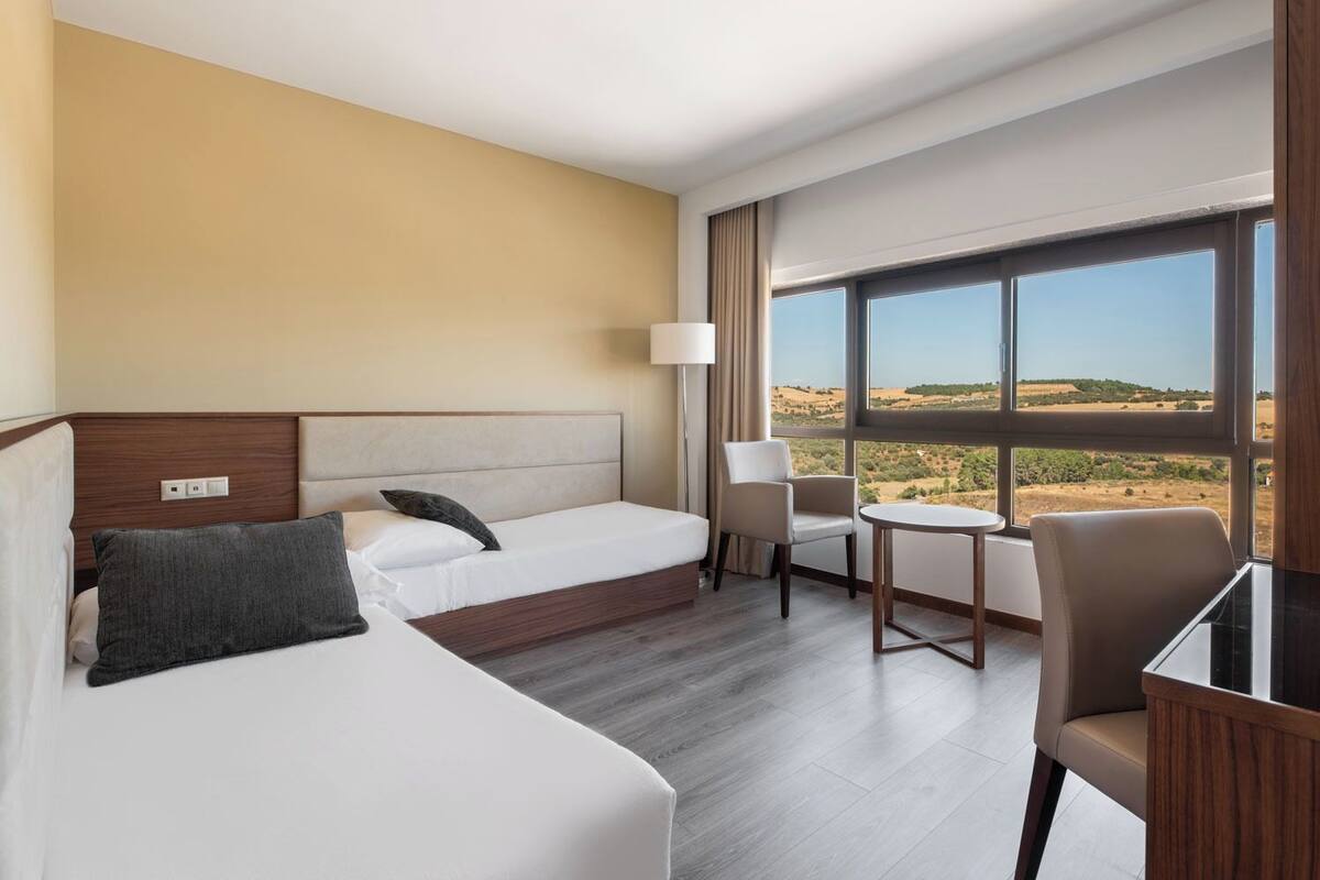 A hotel room with two beds, a desk, a chair, and a large window offering a view of the countryside.