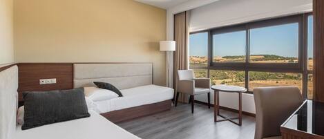 A hotel room with two beds, a desk, a chair, and a large window offering a view of the countryside.