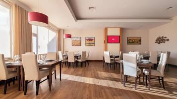 Un vaste espace dédié à la salle à manger, avec de nombreuses tables et chaises, de grandes fenêtres dotées de rideaux et des œuvres d’art accrochées aux murs.