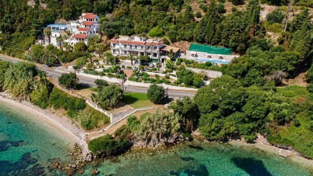 An aerial view of a coastal property with multiple buildings, a beach, and surrounding greenery. - Aurora Beach Hotel (Corfu)