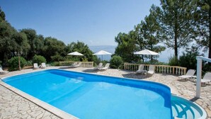 A swimming pool with lounge chairs and umbrellas, surrounded by stone flooring and greenery. - Aurora Beach Hotel (Corfu)