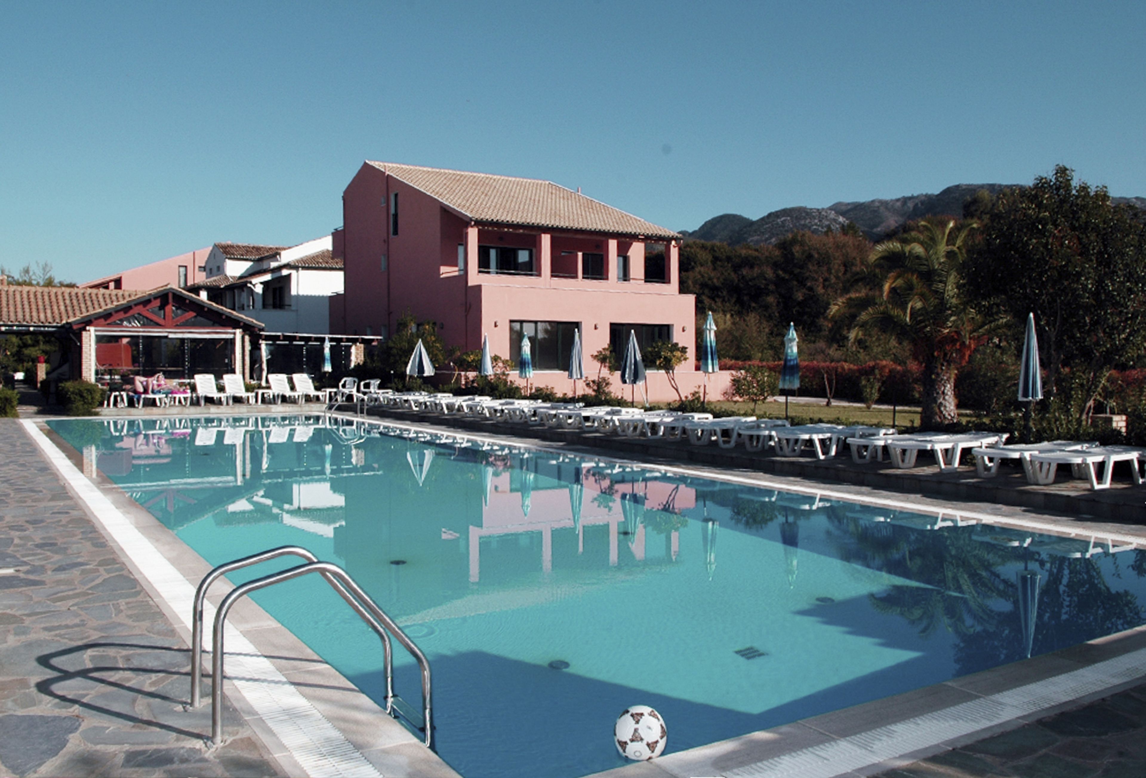 A swimming pool with lounge chairs and umbrellas, a building with a red facade, and a mountainous backdrop.