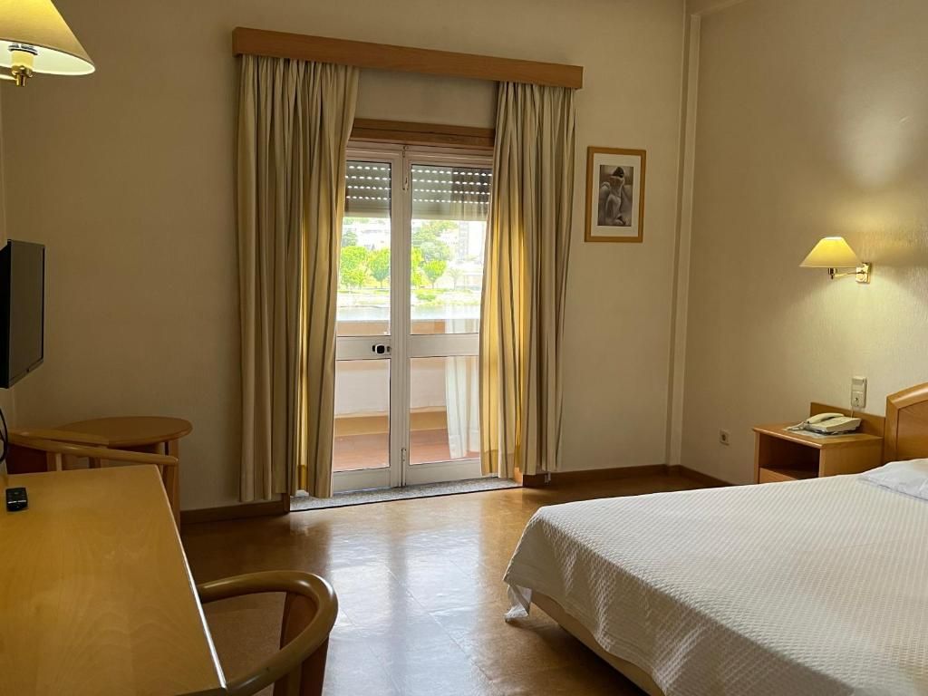 Hotelkamer met een bed, bureau, stoel, nachtkastje, lamp, raam met gordijnen en een deur naar een buitenruimte.