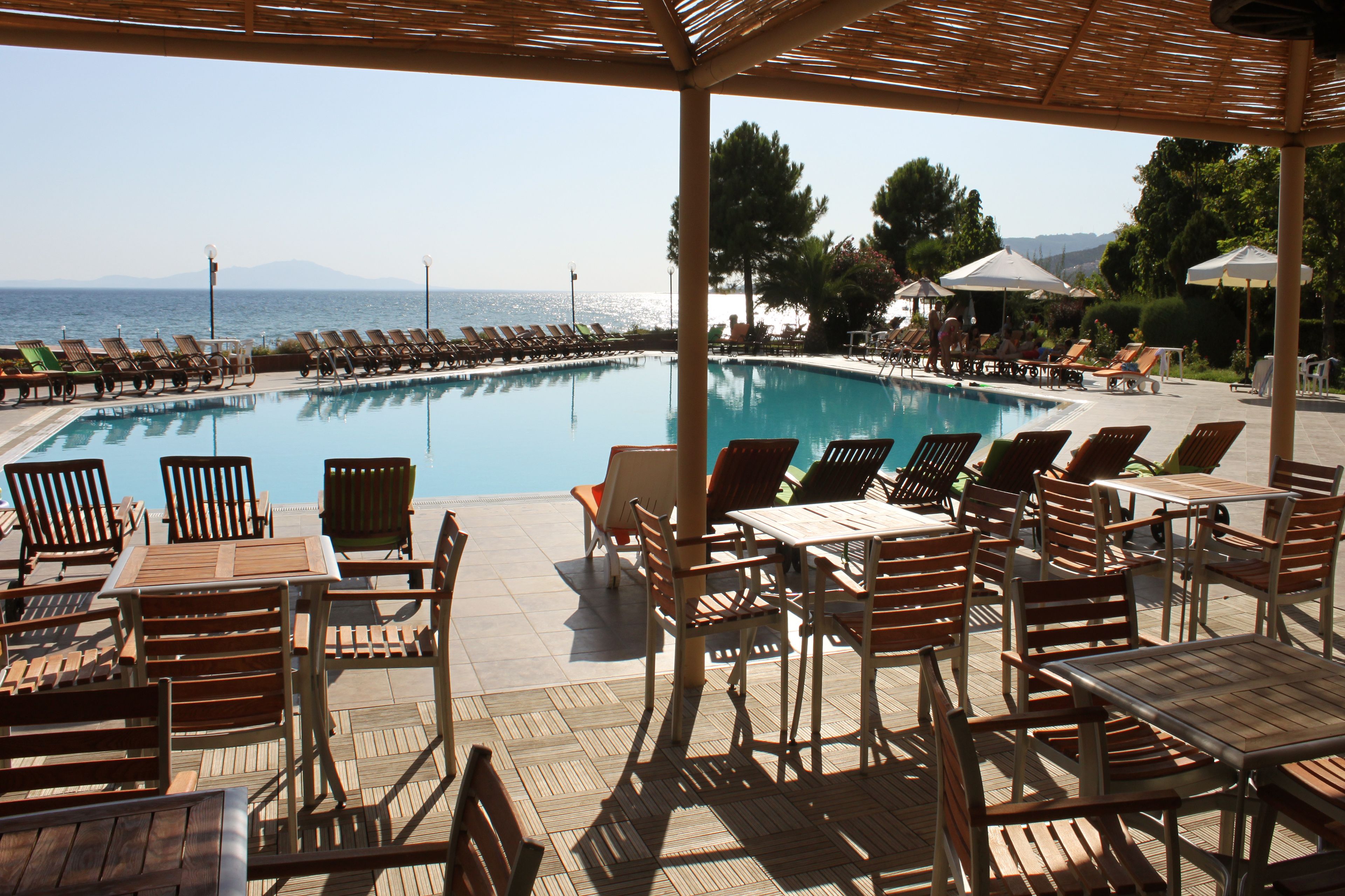 A poolside area with lounge chairs and tables, a clear view of the sea, and a covered seating area.