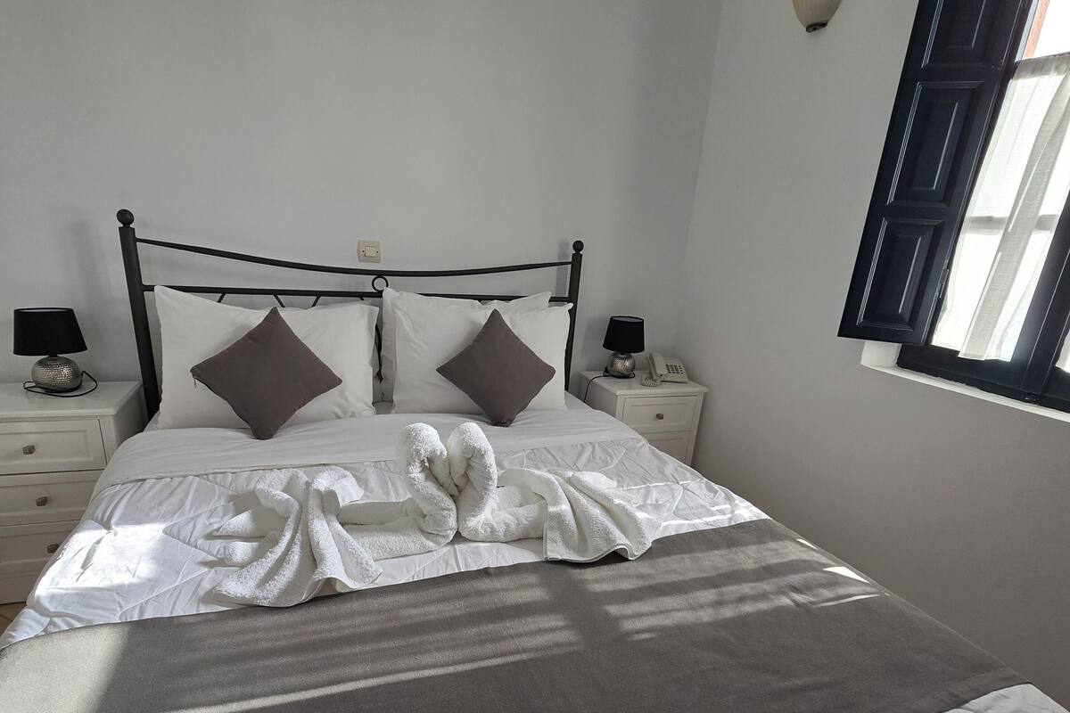 Una cama bien hecha con edredón blanco y dos almohadas, una mesita de noche con lámpara, y una lámpara de pared.