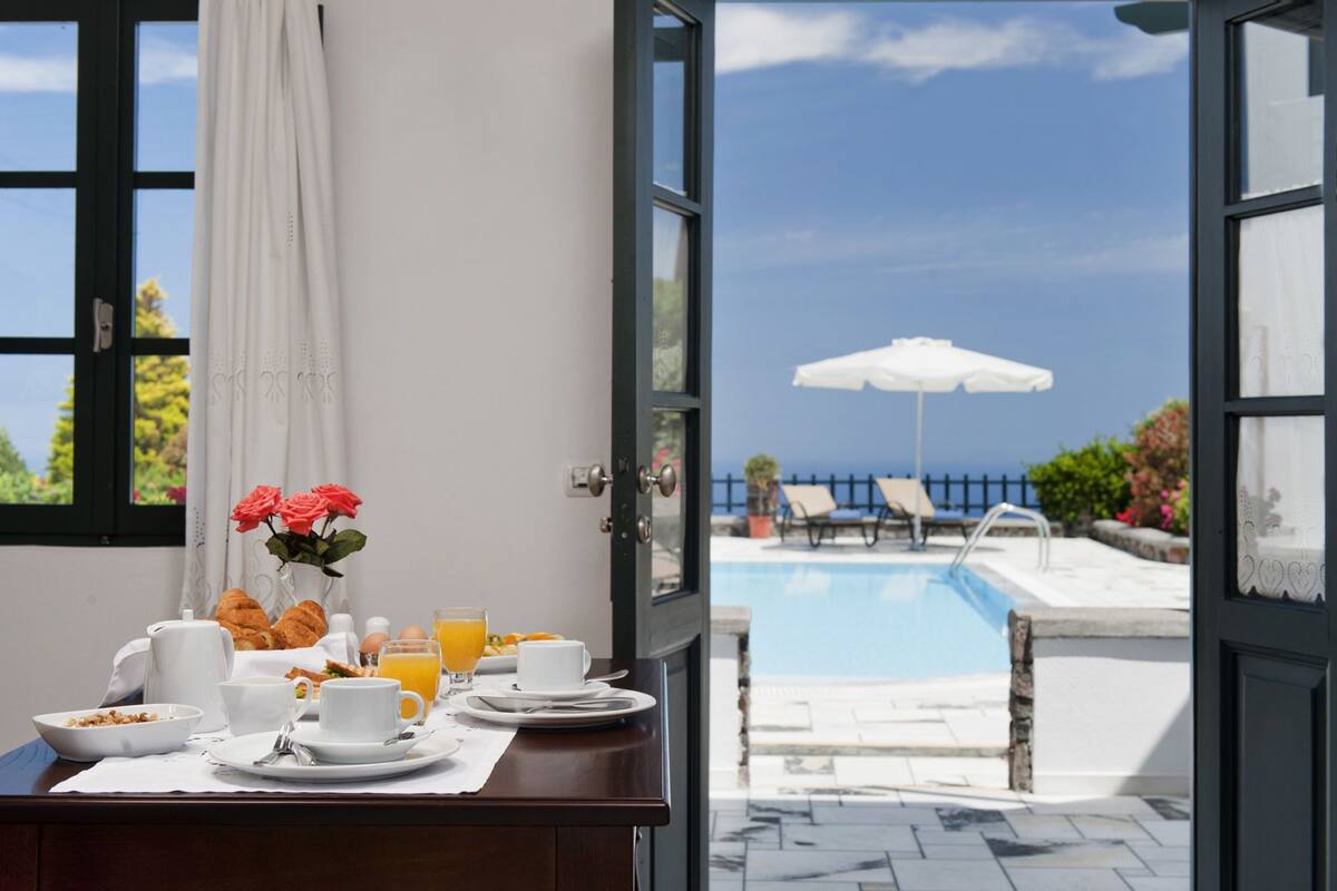 Desayuno con cruasanes, huevos y jugo de naranja sobre una mesa, con vista a una piscina y sillas de descanso con sombrilla.