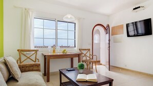 Ein Wohnzimmer mit einer Couch, einem Holztisch mit einem Buch, einer kleinen Topfpflanze und einem Fernseher an der Wand.
