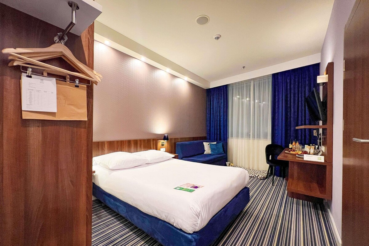 A hotel room with a bed, a desk with a chair, a hanging coat rack, and a window with curtains.