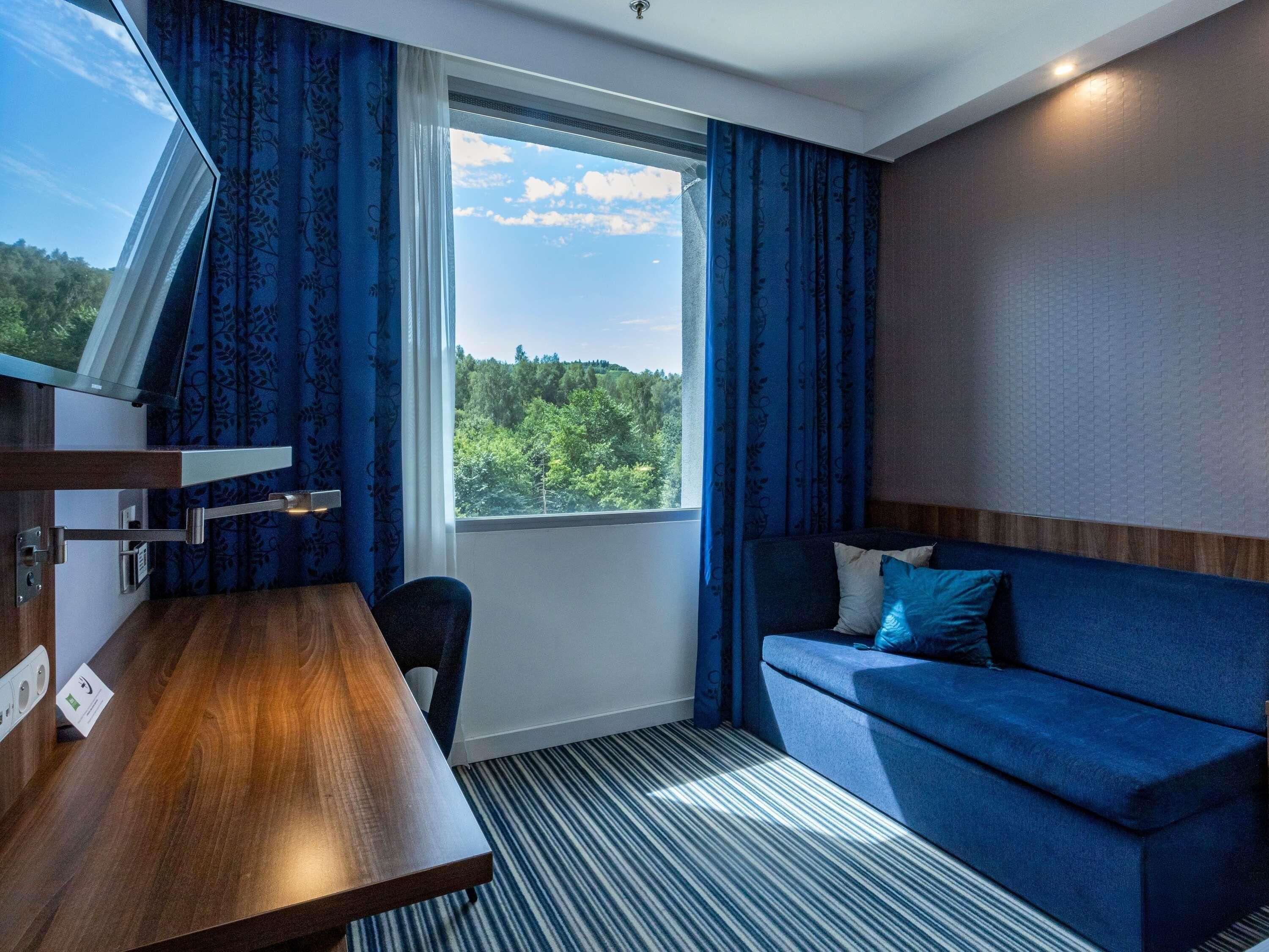 A hotel room with a wooden desk, a sofa, a window with a view of trees, and a striped carpet.