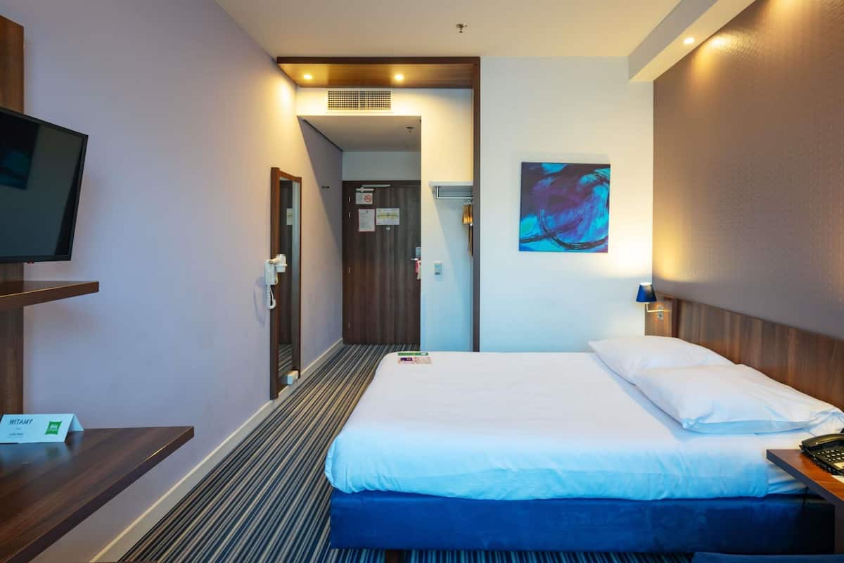 A hotel room with a bed, a desk with a phone, a TV, a painting on the wall, and a door leading to a hallway.