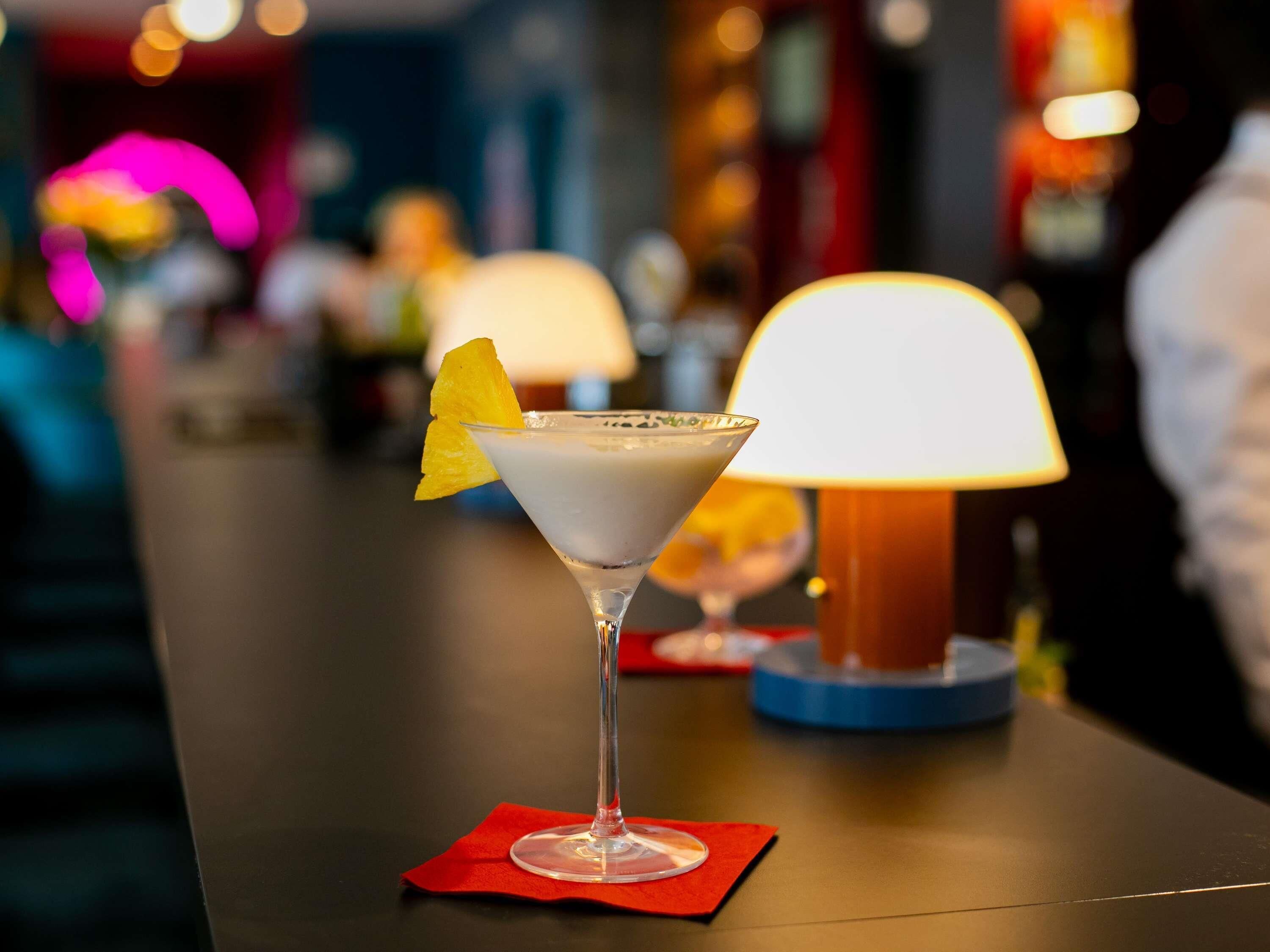 A martini glass with a yellow fruit garnish on a red napkin, placed on a bar counter with a lit lamp in the background.