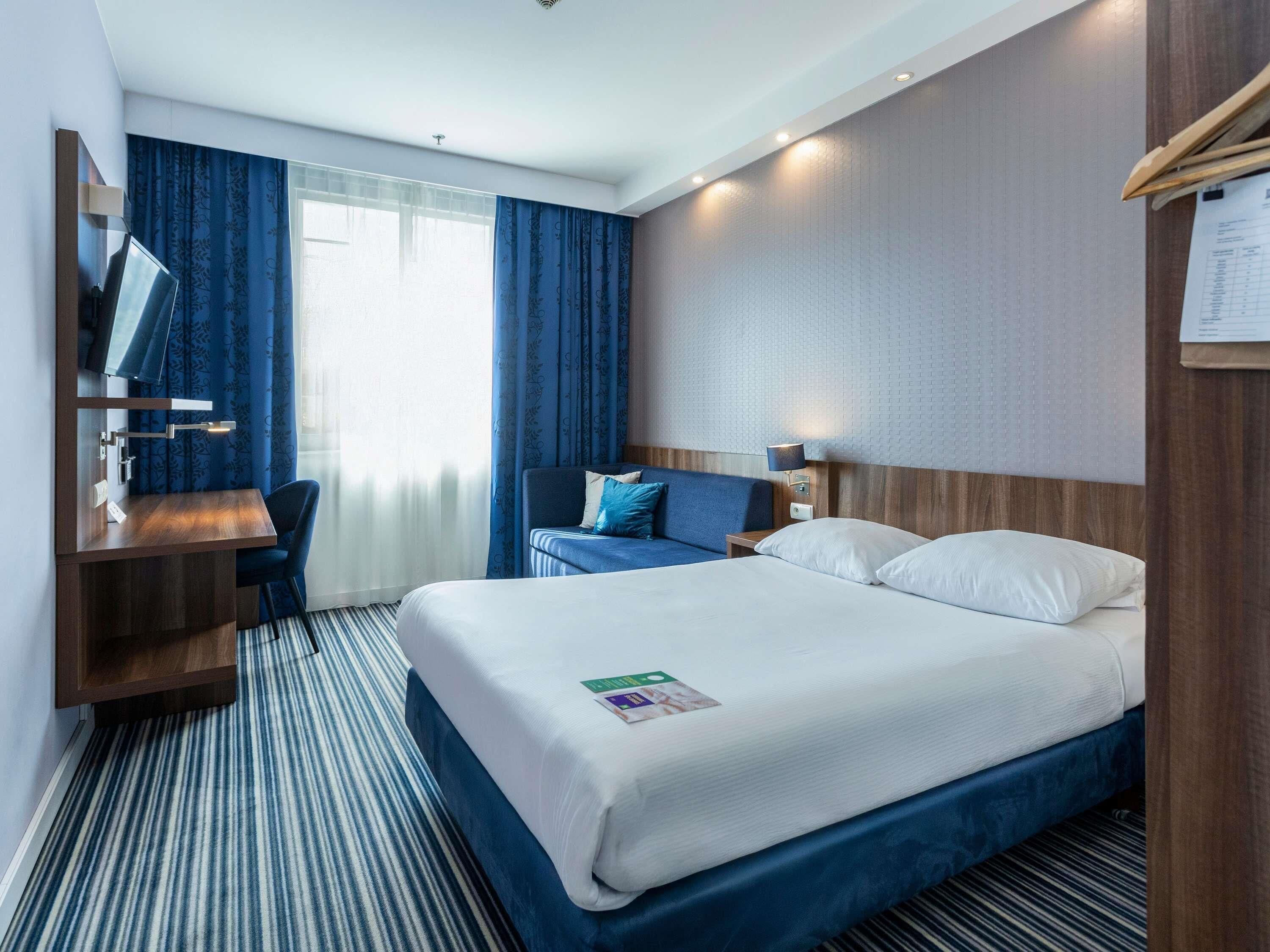 A hotel room with a bed, a desk, a chair, a TV, and a window with curtains.