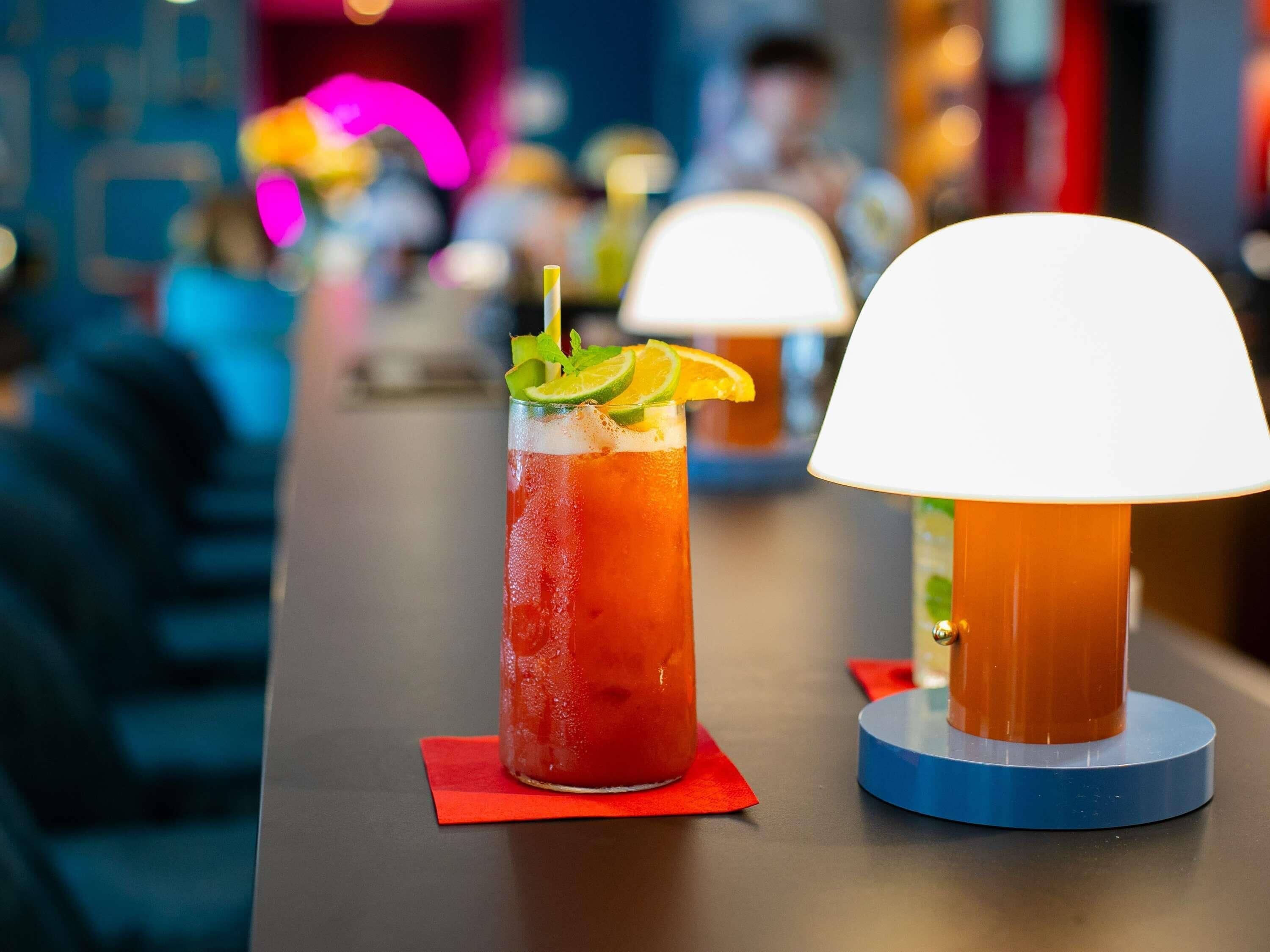 Un bar con un vaso alto de una bebida roja adornado con lima y menta, y una lámpara de mesa con pantalla blanca.