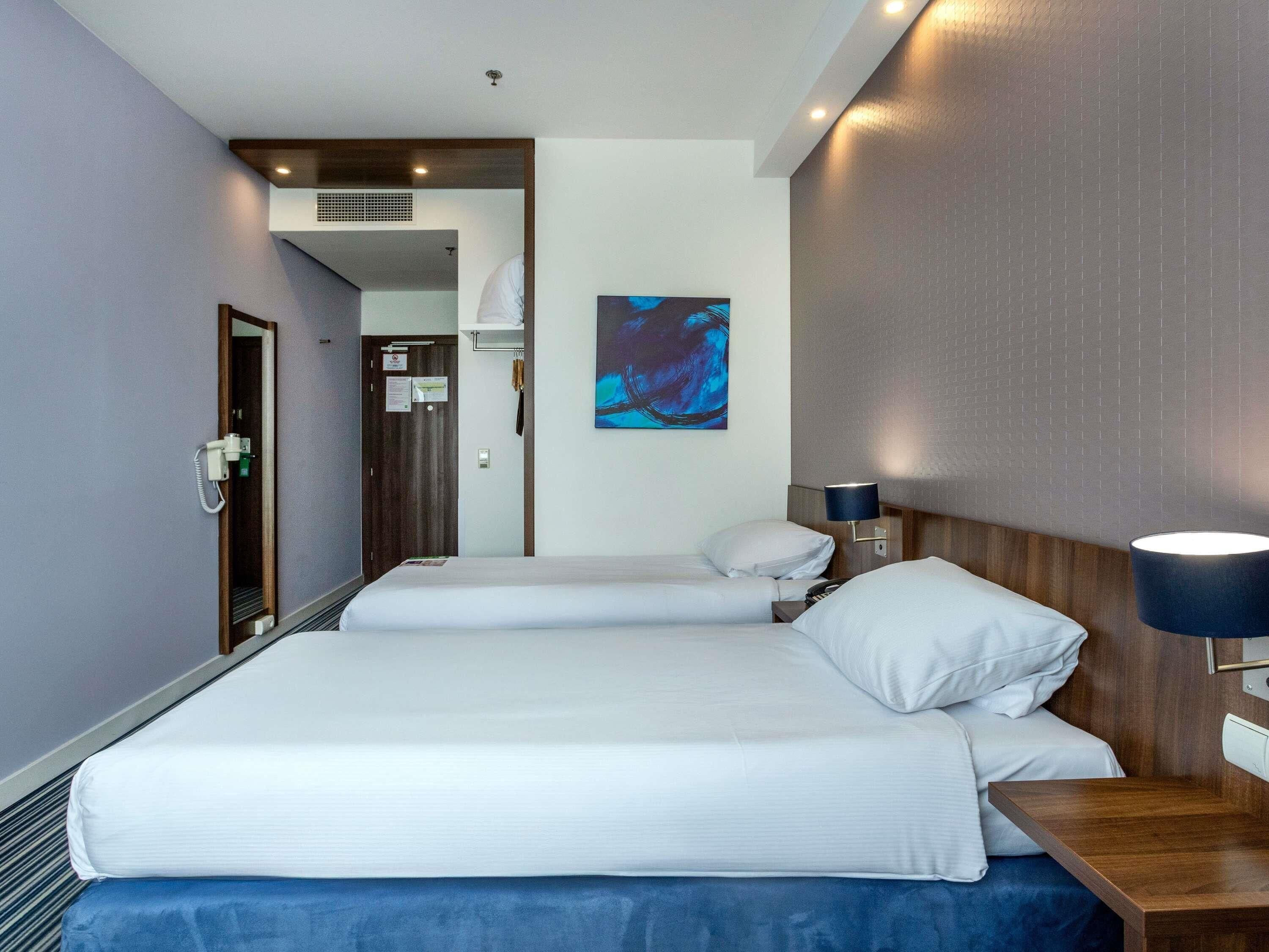 A hotel room with a bed, bedside tables, a lamp, a painting on the wall, and a door leading to another room.