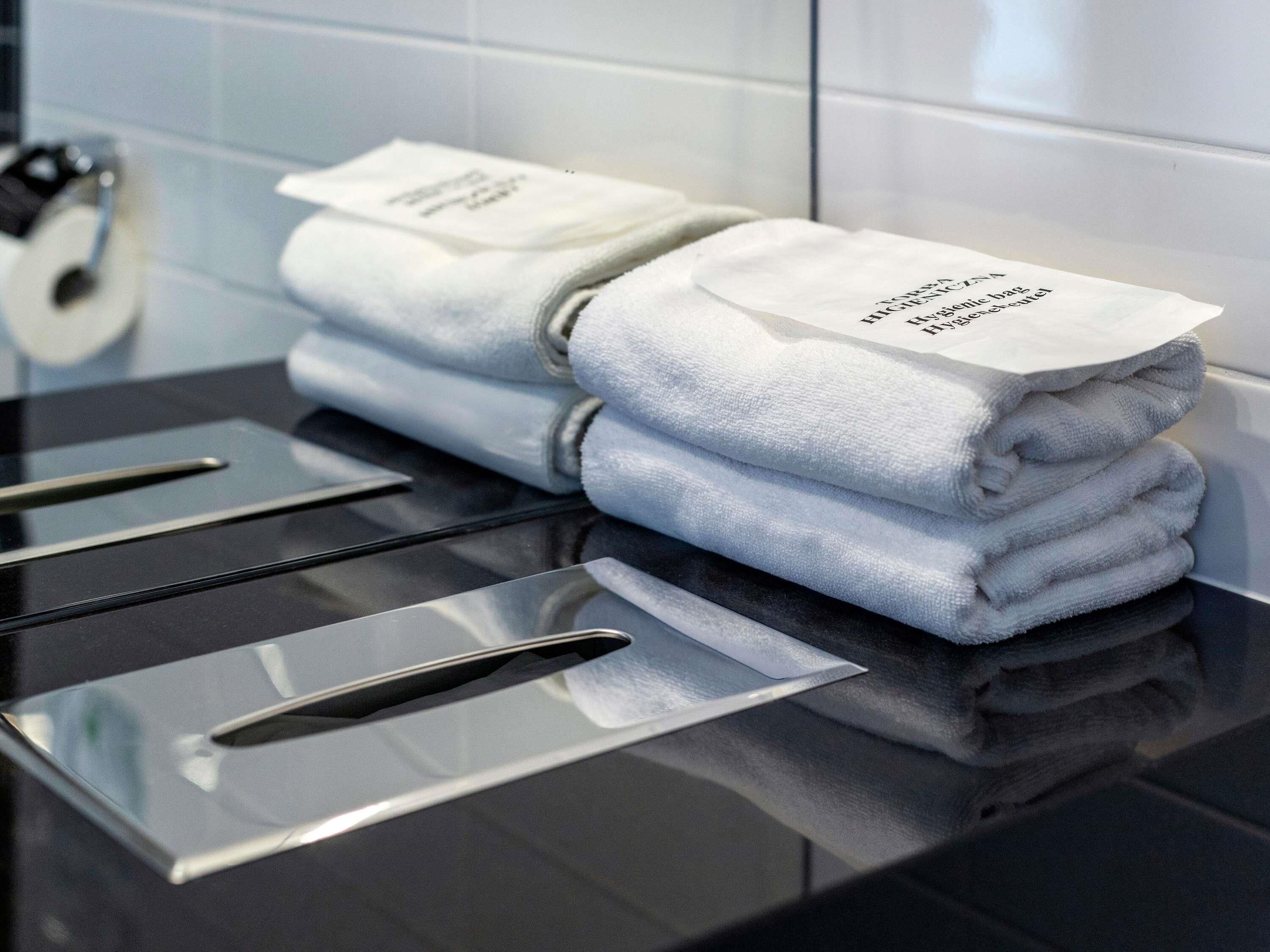 A stack of folded towels with a label on top, placed on a black surface with a metallic towel rack in a bathroom setting.