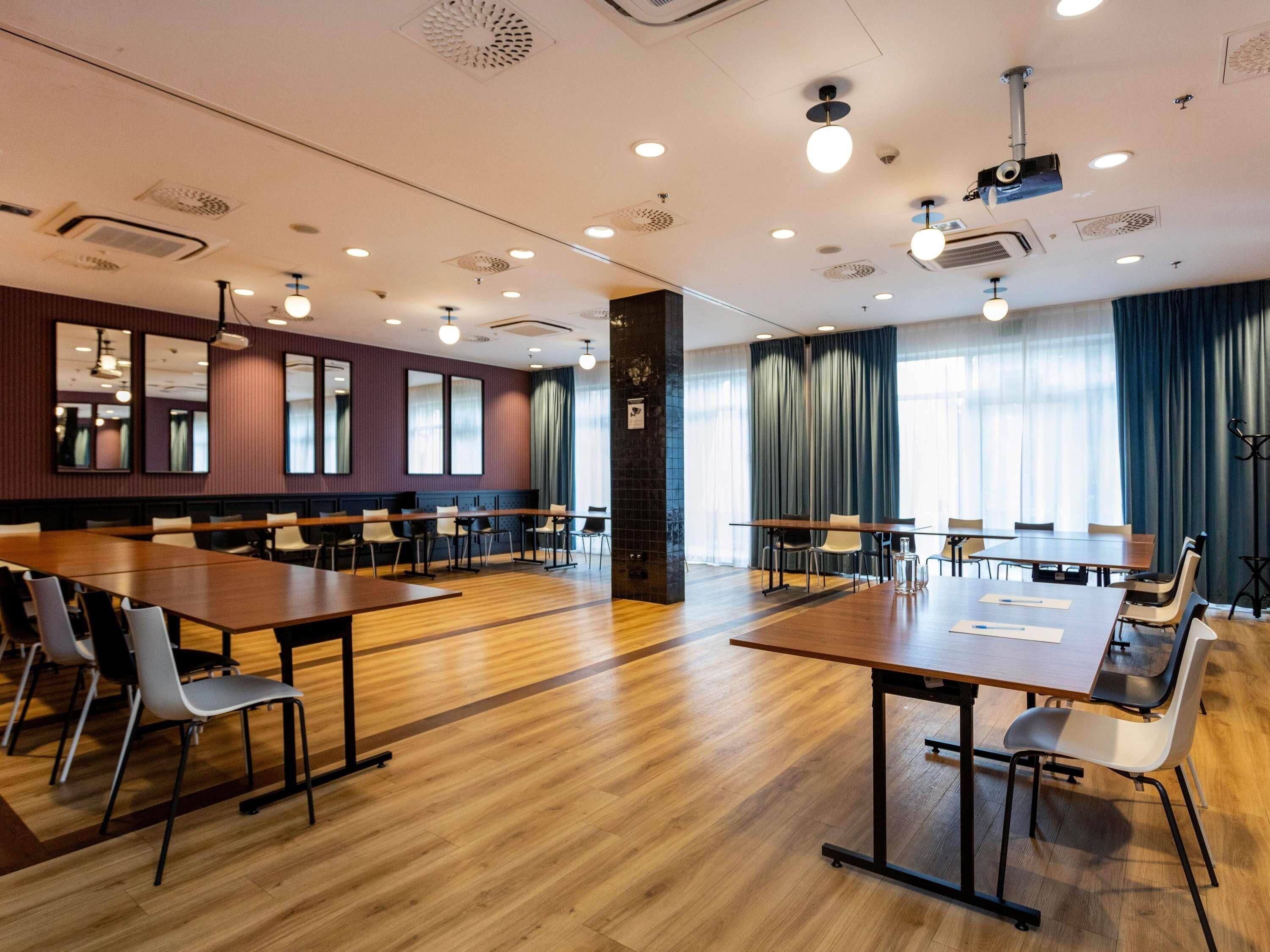 A conference room with multiple tables and chairs, a large window with curtains, and a ceiling with recessed lighting and ventilation.