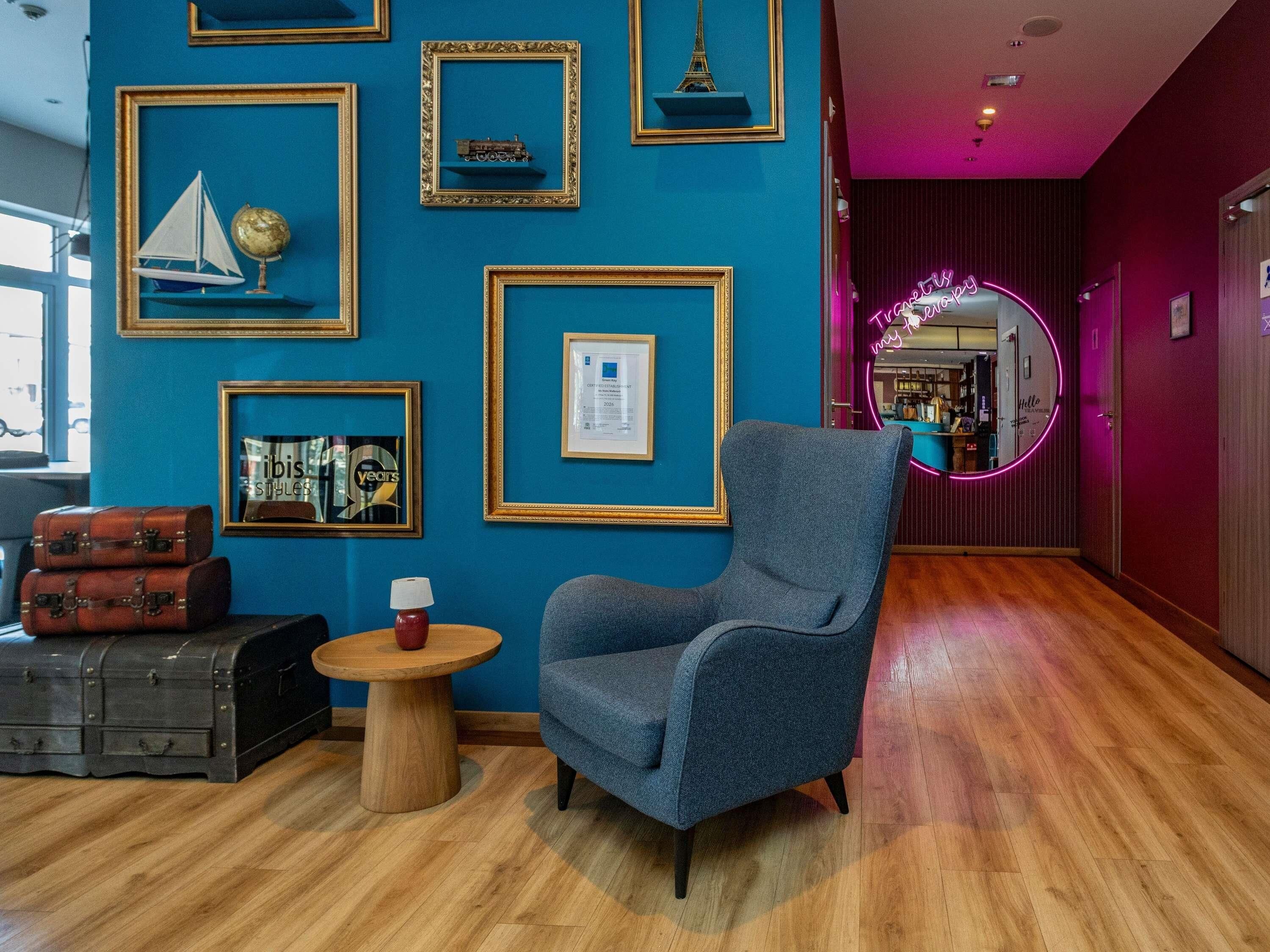 Una habitación con pared azul, cuadros enmarcados, un sillón azul, una mesa de madera y una lámpara roja.