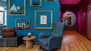 Une pièce avec un mur bleu, des tableaux encadrés, un fauteuil bleu, une table en bois et une lampe rouge.