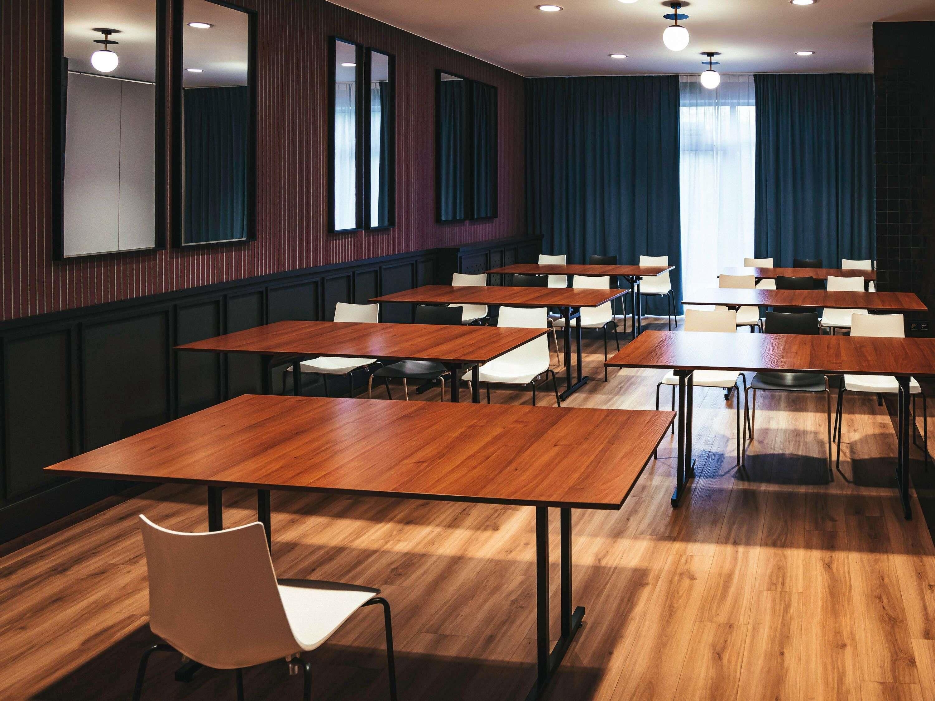 A conference room with multiple tables and chairs, wooden flooring, and large windows with curtains.