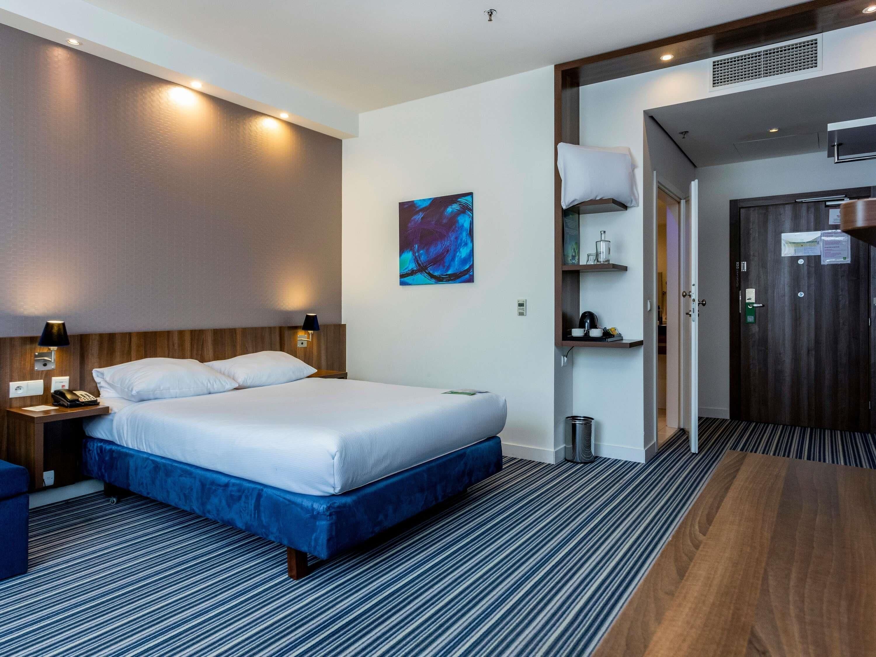 A hotel room with a bed, bedside table, lamp, artwork, and a door leading to a bathroom.